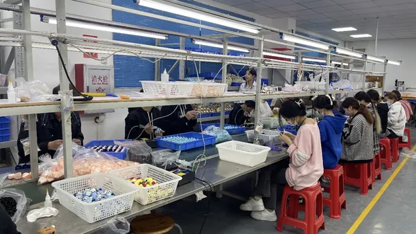 Operators on both sides of a bench handle small coloured parts, with sorting trays and tools on the work surface.
